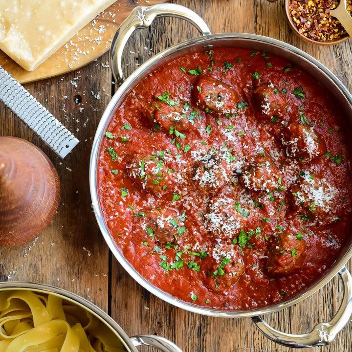 A CopperBond Copper Induction Soup Pot, 3-Quart, filled with meatballs in tomato sauce, garnished with parsley and grated cheese on a wooden table next to a cheese block, grater, chili flakes, pepper mill, and pasta. Handcrafted in Italy and induction compatible.