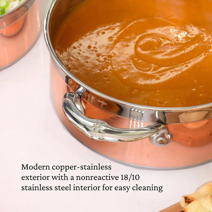 A CopperBond Copper Induction Soup Pot, 3-quart, boasting a polished copper exterior and nonreactive 18/10 stainless steel interior, handcrafted in Italy and induction compatible, filled with sauce. Nearby vegetables and a wooden spoon complete the elegant scene.