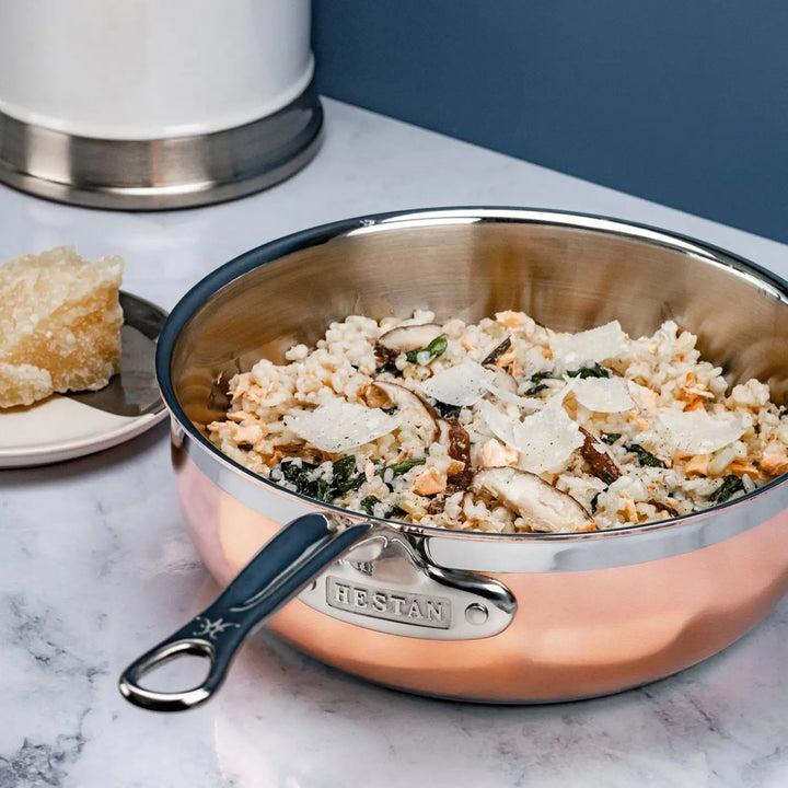 A CopperBond Copper Induction Essential Pan, expertly handcrafted in Italy, holds risotto topped with mushrooms, greens, and cheese shavings. A cheese wedge sits on a small plate nearby as the pan rests elegantly on the marble countertop.