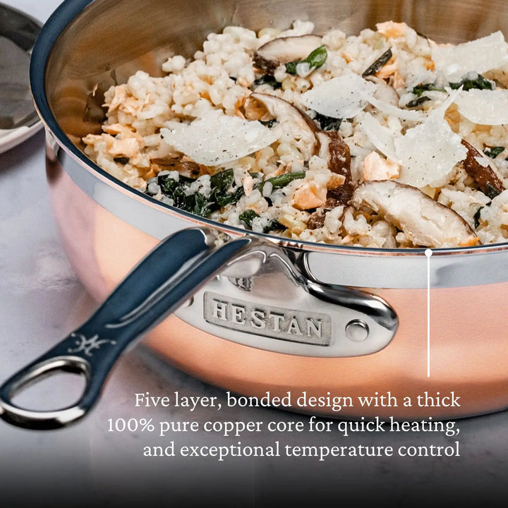 A Copper Induction Essential Pan by CopperBond, handcrafted in Italy, features HESTAN on the handle. The five-layer stainless steel with a copper core ensures quick heating and precise control. It contains a dish with rice, mushrooms, spinach, and cheese shavings.