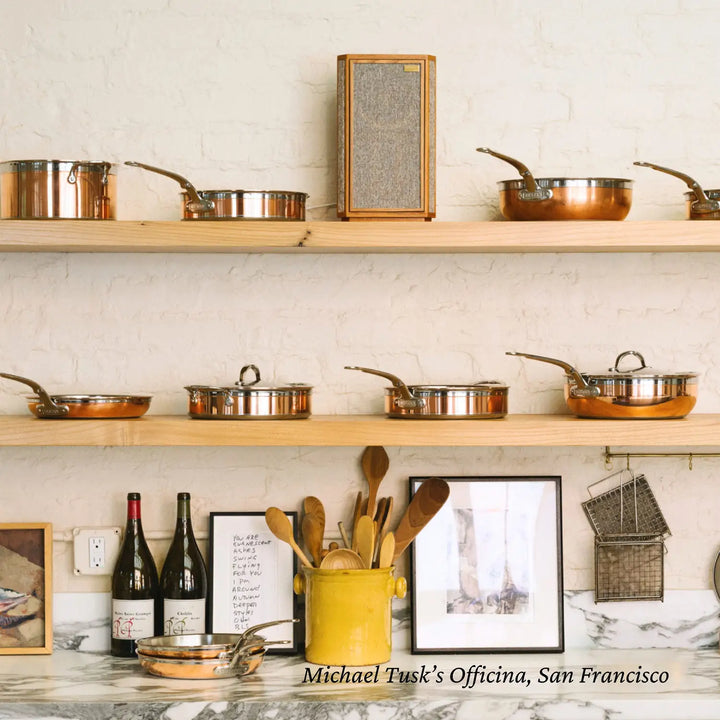 A kitchen with wooden shelves showcases CopperBonds Copper Induction Essential Pans. Below, wine bottles, framed art, and wooden utensils sit on a marble countertop against a white brick wall. Text reads: Michael Tusk’s Officina, San Francisco. Handcrafted in Italy.