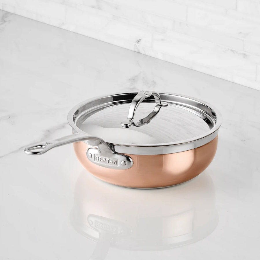 A CopperBond Copper Induction Essential Pan with a stainless steel handle and glass lid sits on a white marble countertop by a white tiled wall. This pan is oven safe to 600°F, offering versatile cooking options.