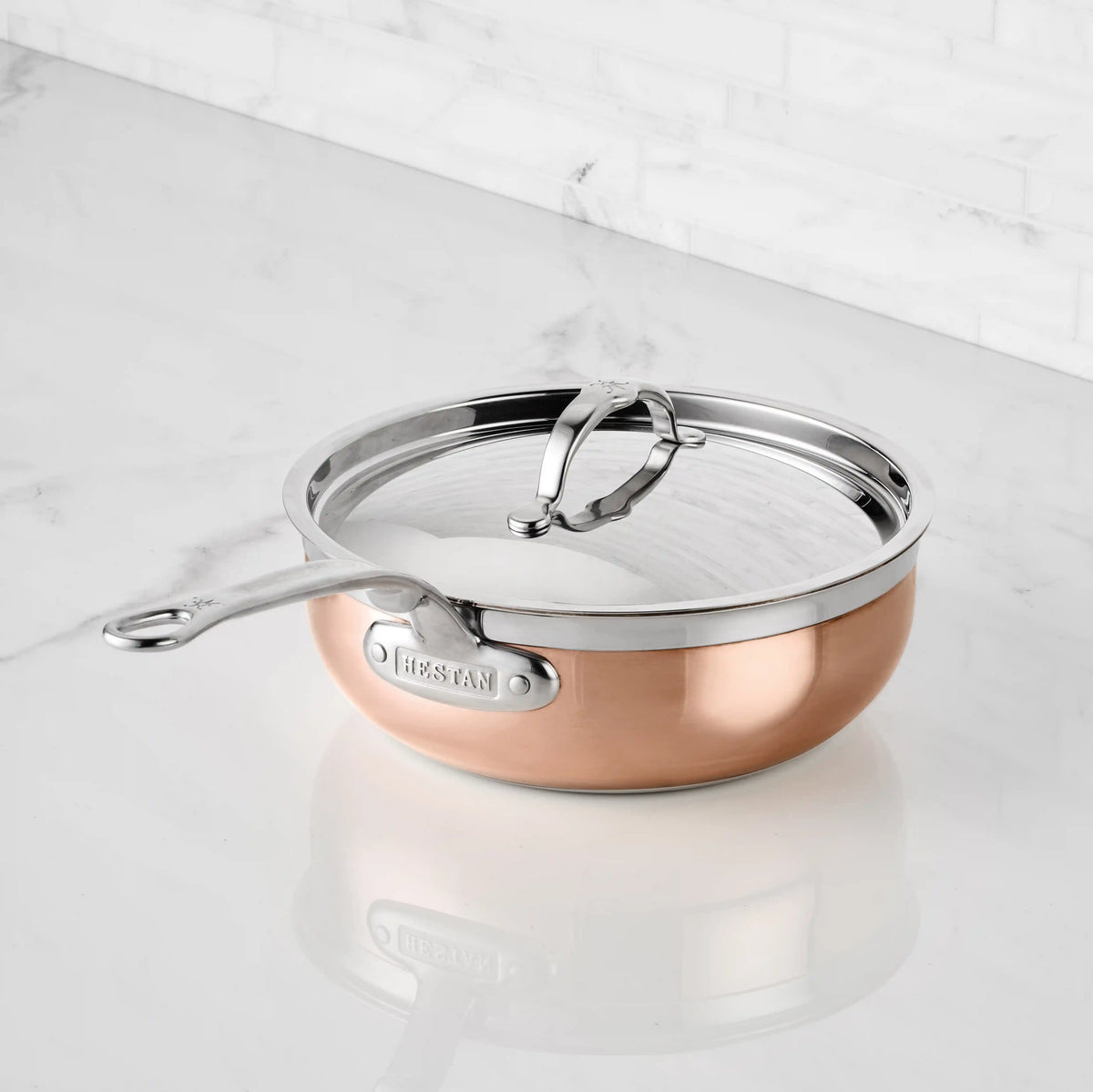 A CopperBond Copper Induction Essential Pan with a stainless steel handle and glass lid sits on a white marble countertop by a white tiled wall. This pan is oven safe to 600°F, offering versatile cooking options.