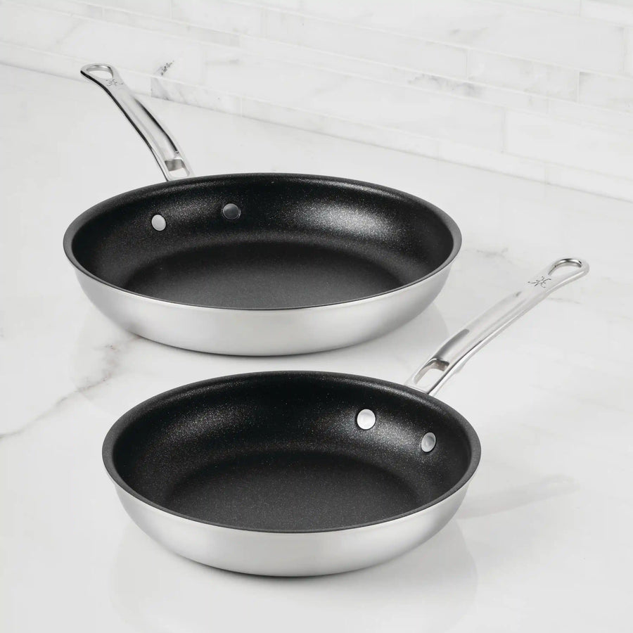 Two Thomas Keller Insignia Commercial Clad Stainless Steel TITUM® Nonstick Skillets, one large and one small, with overlapping black nonstick interiors and sleek metal handles, sit on a white marble countertop.