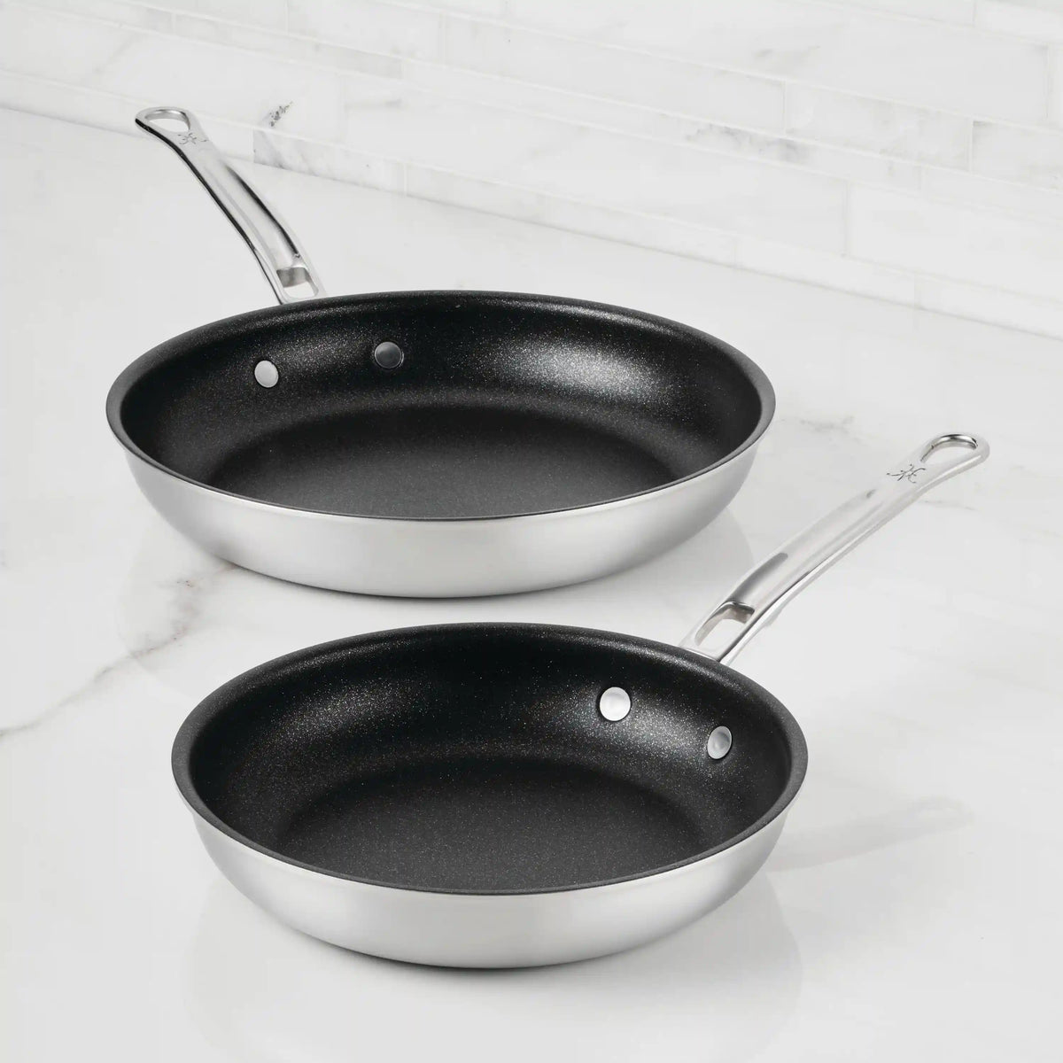 Two Thomas Keller Commercial Clad Stainless Steel TITUM® Nonstick Skillets with black interiors rest on a white marble countertop, offering professional performance and long metal handles—ideal for any kitchen.