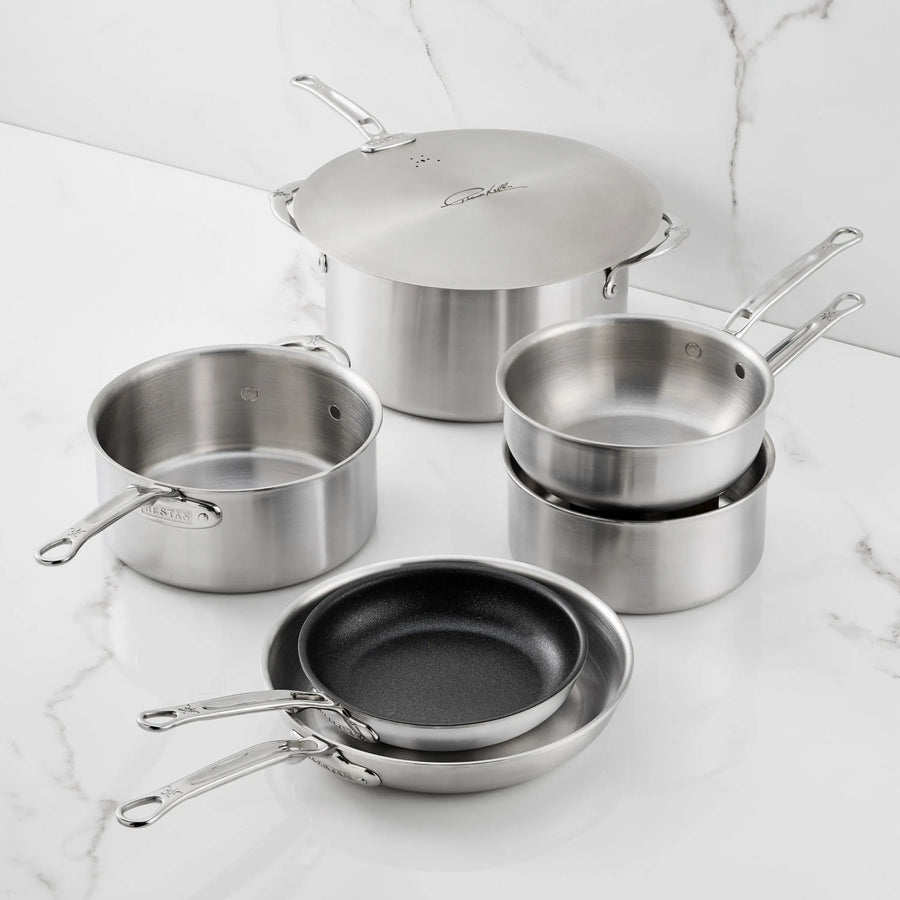 The Thomas Keller Commercial Clad Stainless Steel 7-Piece Cookware Set, featuring saucepans, pots with lids, and nonstick frying pans, is arranged on a white marble countertop for professional culinary performance.