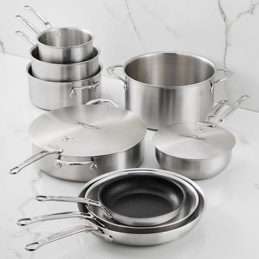 The Thomas Keller Commercial Clad Stainless Steel 11-Piece Cookware Set features saucepans with lids, a stock pot, frying pans, and sauté pans—all designed for professional results and shown neatly arranged on a white marble countertop.