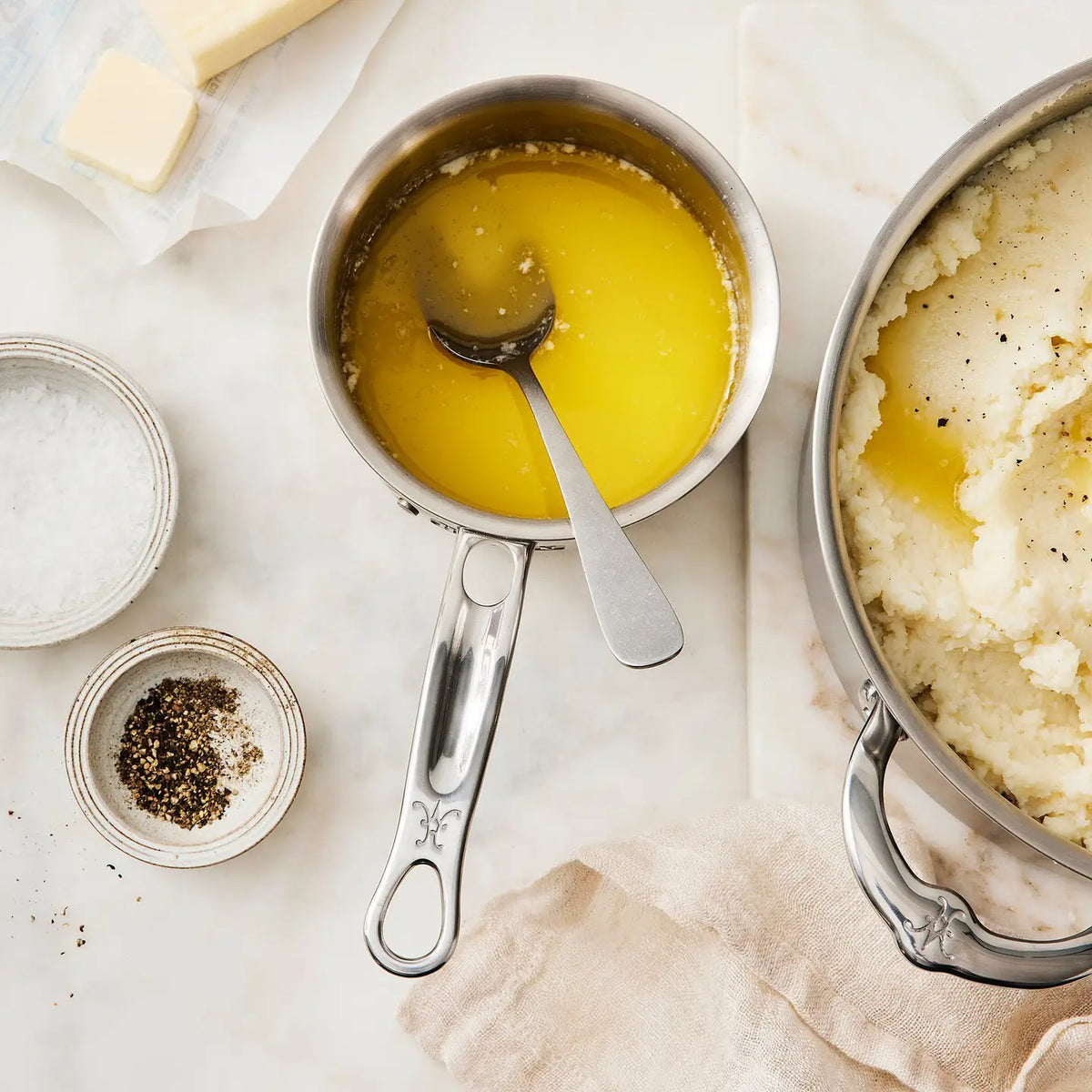 A Thomas Keller Commercial Clad Stainless Steel 0.75-Quart Butter Warmer filled with melted butter and a spoon is next to mashed potatoes, with salt, pepper bowls, a butter stick, and a light cloth on a marble countertop.