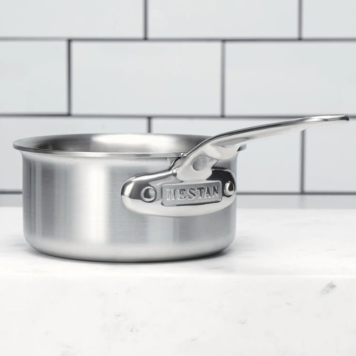 A Thomas Keller Insignia Commercial Clad Stainless Steel 0.75-Quart Butter Warmer with a long handle sits on a white marble surface, its polished finish gleaming against the white tiled wall. Perfect for any culinary adventure, this cookware combines style and durability seamlessly.