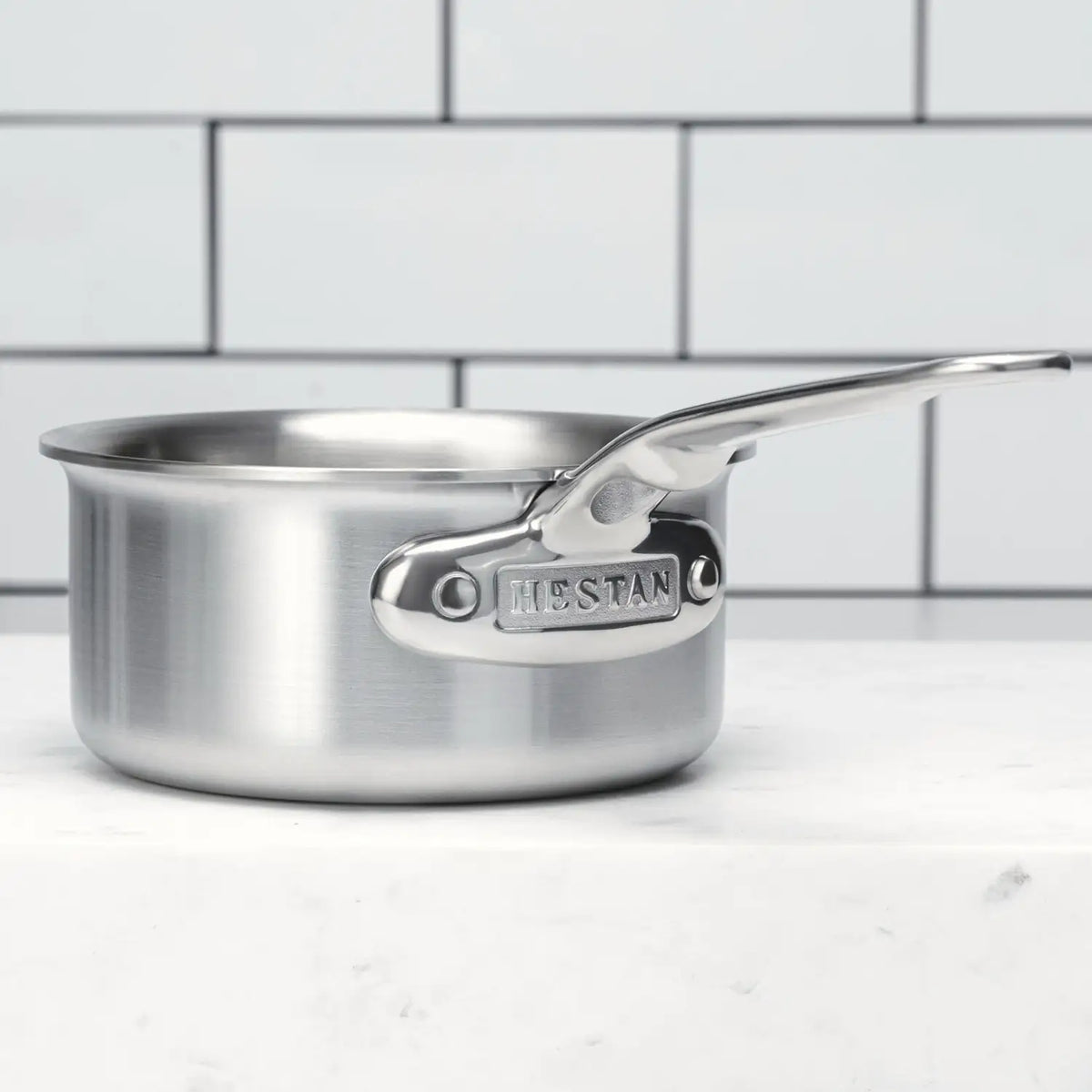 The Thomas Keller Commercial Clad Stainless Steel 0.75-Quart Butter Warmer rests on a white surface before a white tile wall, with the word "Hestan" engraved on its handle attachment.