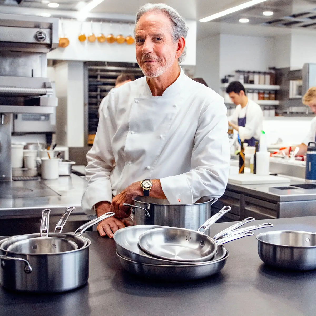 A chef in a white coat stands in a modern kitchen behind a counter displaying Thomas Keller Commercial Clad tri-ply cookware, including the Stainless Steel 0.75-Quart Butter Warmer, with kitchen staff working in the background.