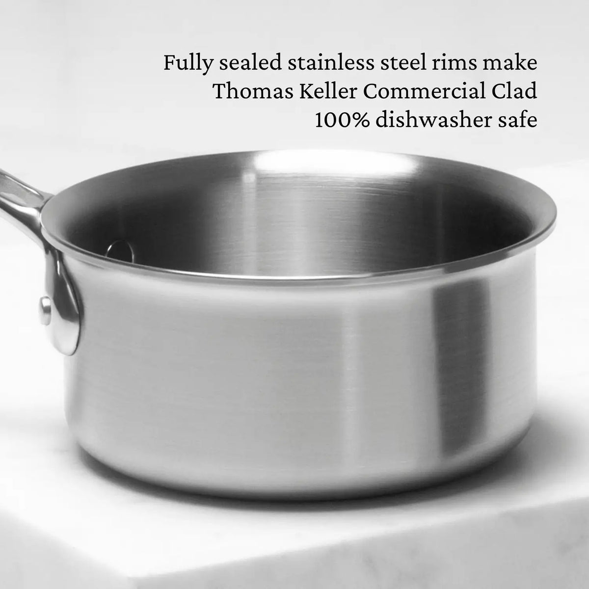 A Thomas Keller Commercial Clad 0.75-quart stainless steel butter warmer with handle sits on a white countertop. Text above reads: Fully sealed rims make it 100% dishwasher safe chef-designed tri-ply cookware.