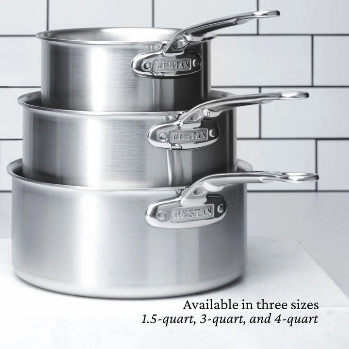 On a white countertop, three Thomas Keller Insignia Commercial Clad Stainless Steel Saucepans with polished handles and engraved branding are stacked. A tile backsplash is visible, and text indicates availability in 1.5-quart, 3-quart, and 4-quart sizes.