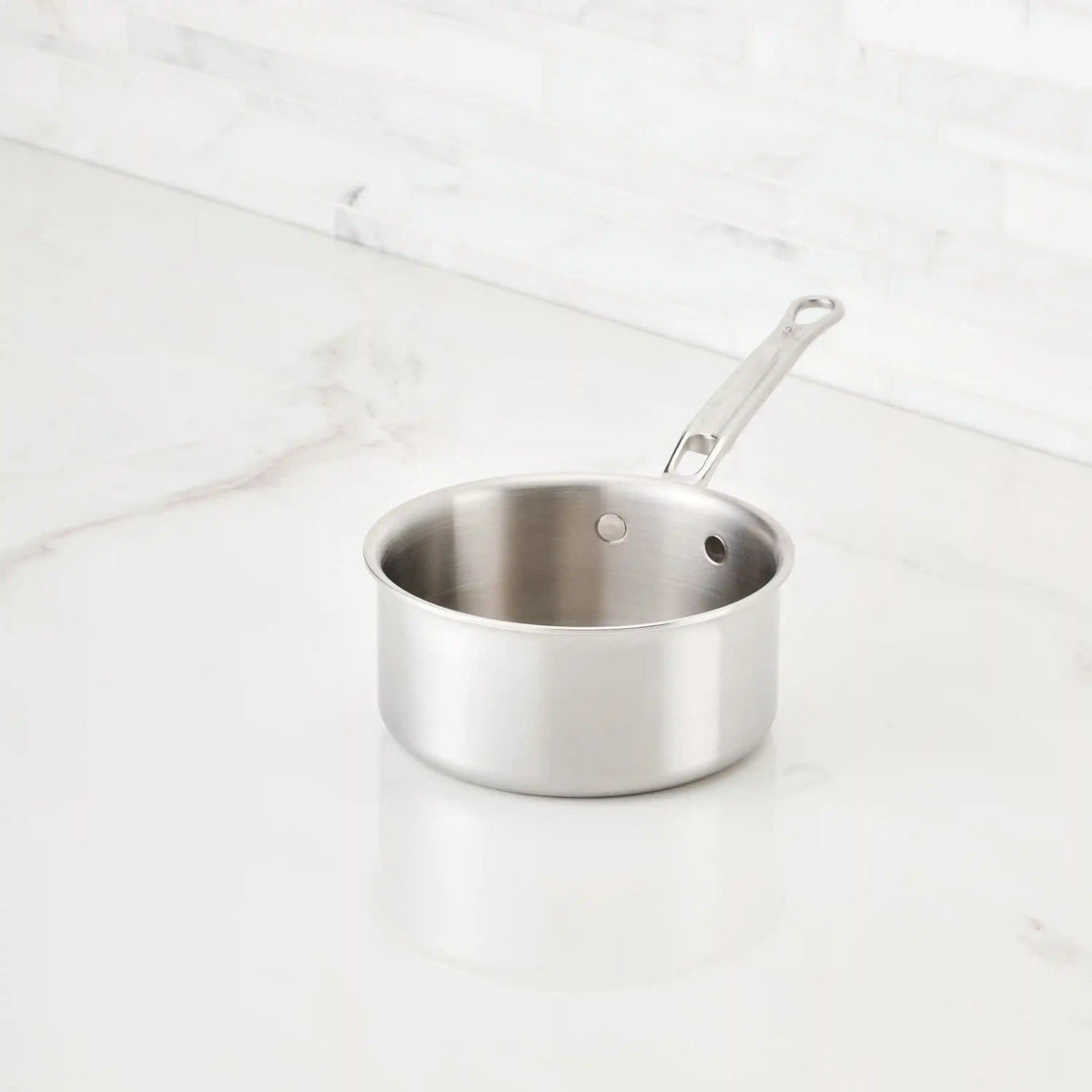 The Thomas Keller Commercial Clad Stainless Steel Saucepan, featuring tri-ply construction and a handle, sits on a white marble countertop in a kitchen with a white tiled backsplash, delivering professional performance.