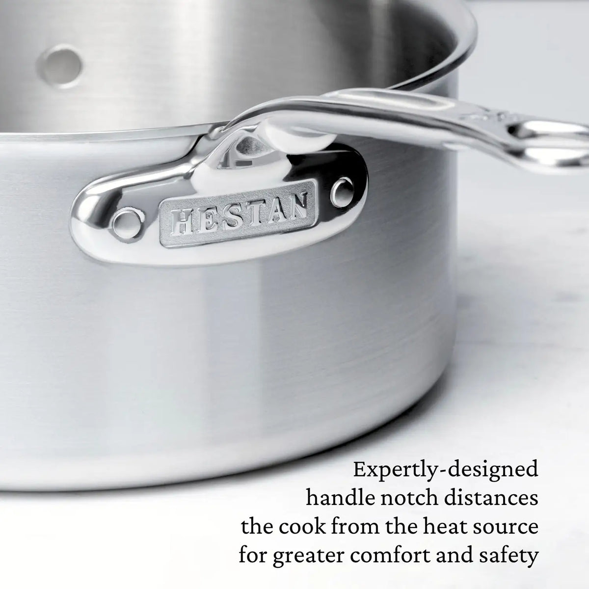 Close-up of a Thomas Keller Commercial Clad Stainless Steel Saucepan features the brand name engraved on the handle and a notched design. Tri-ply construction enhances comfort and safety for professional culinary performance.