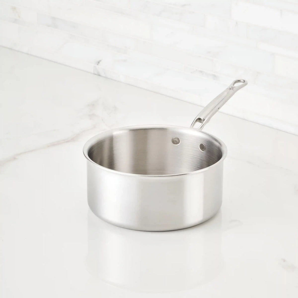 The Thomas Keller Commercial Clad Stainless Steel Saucepan, featuring tri-ply construction and a long handle, enhances kitchen performance and style on a white marble countertop with a white tile backsplash.