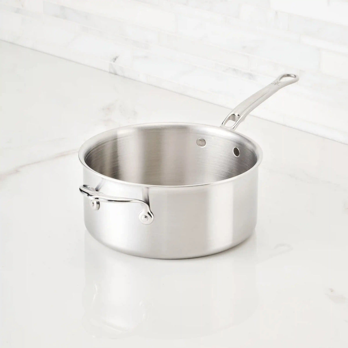 A Thomas Keller Commercial Clad Stainless Steel Saucepan with a long handle gleams on a white marble countertop, its polished tri-ply construction showcasing professional culinary quality against the tiled white backsplash.