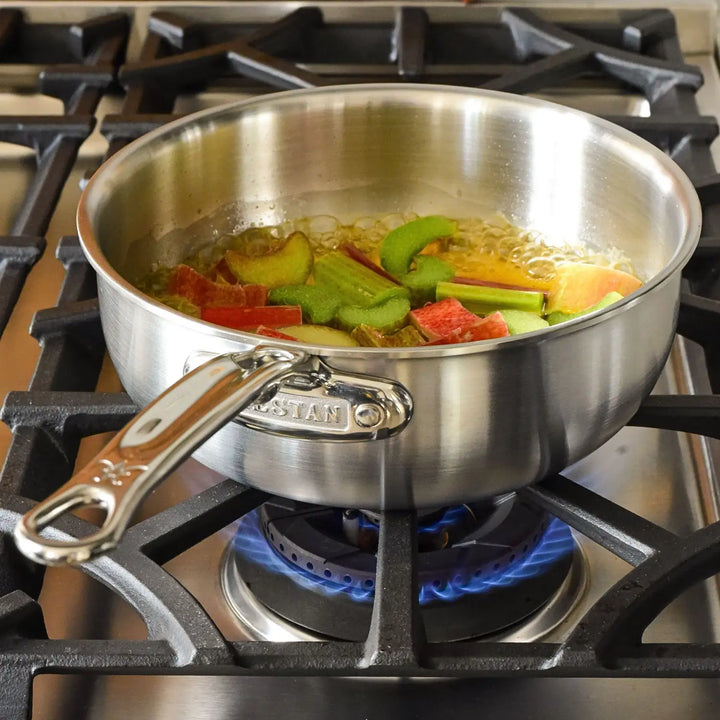 On a lit gas stove burner, the Thomas Keller Insignia Commercial Clad Stainless Steel 2-Quart Saucier demonstrates professional culinary performance with red and green rhubarb simmering inside, showcasing excellent heat conductivity while other burners await their turn.