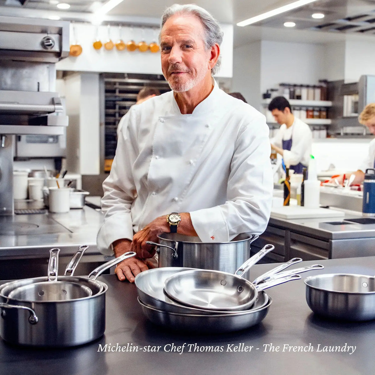 Michelin-star chef Thomas Keller stands in a modern kitchen behind a counter featuring the Thomas Keller Commercial Clad Stainless Steel 2-Quart Saucier. The clean, organized kitchen has staff working in the background. Text: "The French Laundry.