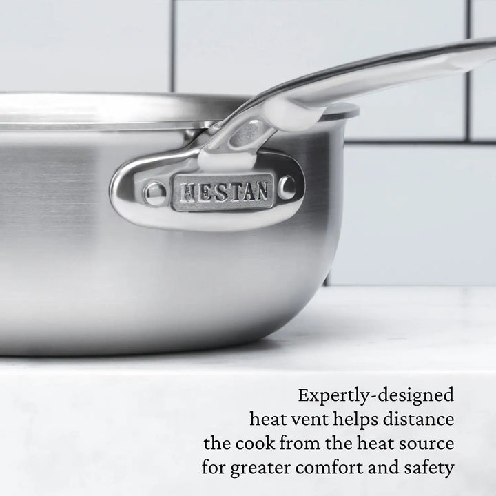 A close-up of a Thomas Keller Insignia 2-quart stainless steel saucier with a shiny handle is set against tilled walls. It features an expertly-designed heat vent that ensures professional culinary performance by keeping the cook at a safe and comfortable distance from the heat source.