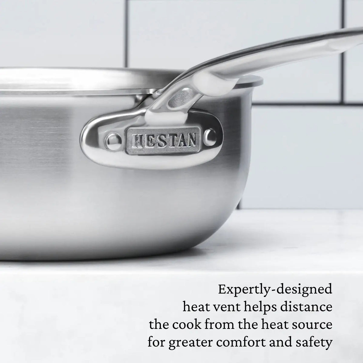 Close-up of a Thomas Keller Commercial Clad Stainless Steel 2-Quart Saucier with HEFTAN engraved on the handle, set against tiles. Text below notes its tri-ply clad design for comfort, safety, and professional-grade culinary performance.