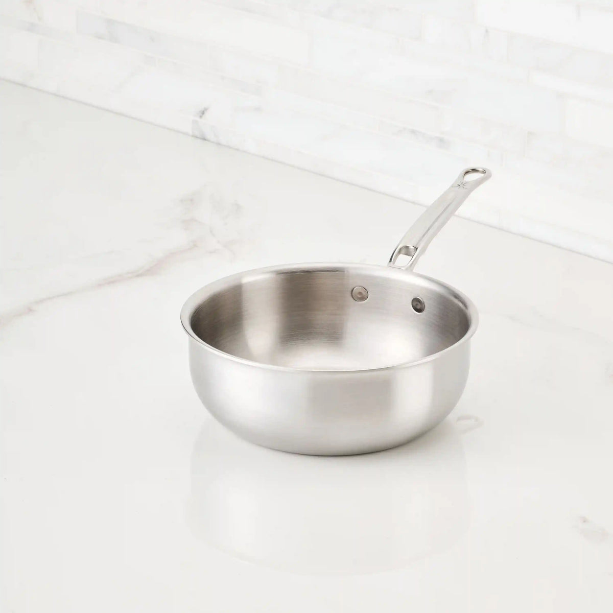 A Thomas Keller Commercial Clad Stainless Steel 11-Piece Cookware Set saucepan sits on a white marble countertop in a kitchen with a light-colored tile backsplash.