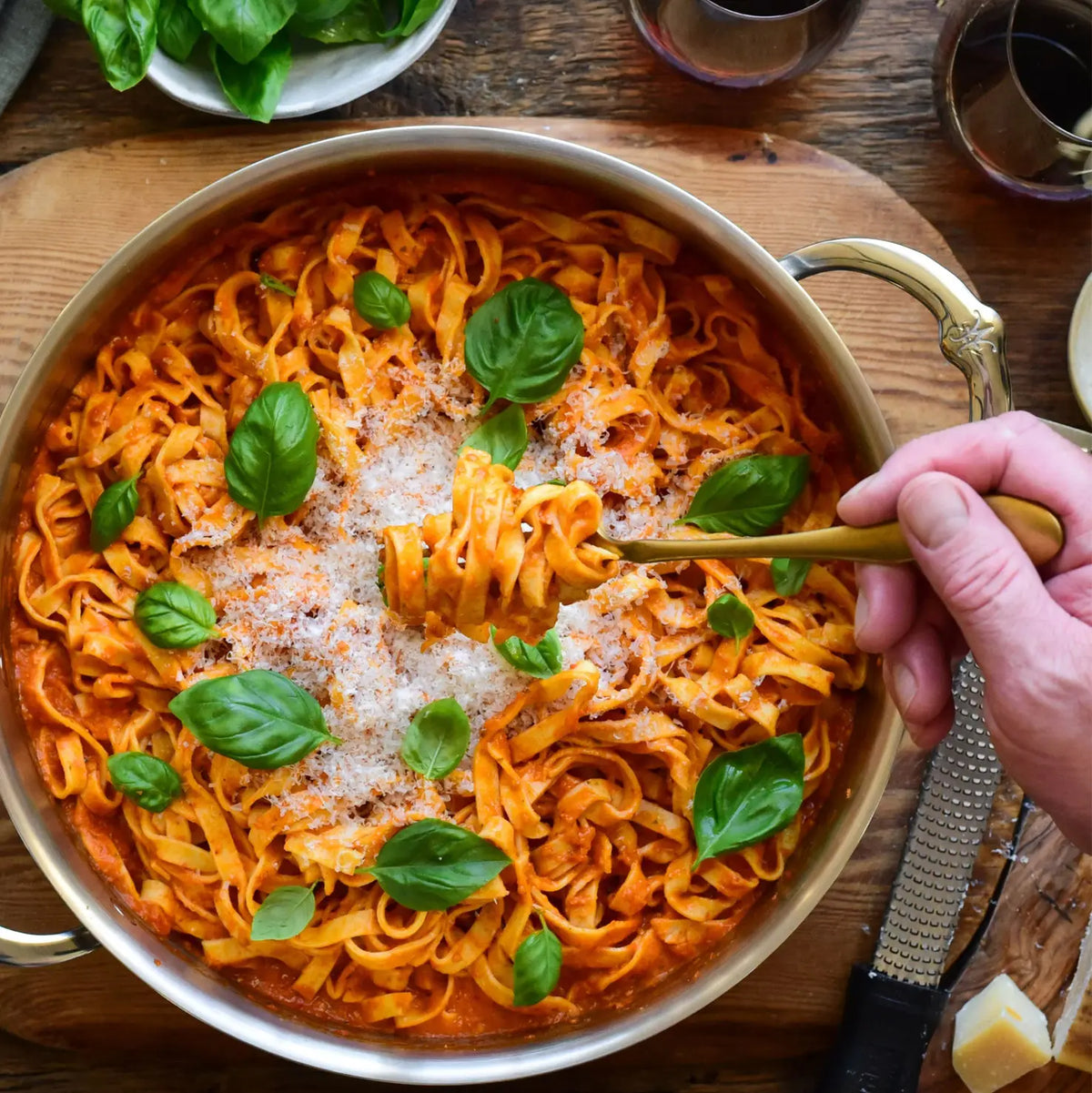 A hand twirls tagliatelle with tomato sauce, cheese, and basil in a Thomas Keller Commercial Clad Stainless Steel Stock Pot on a wooden table, surrounded by fresh basil, cheese, a grater, and red wine for a professional culinary touch.