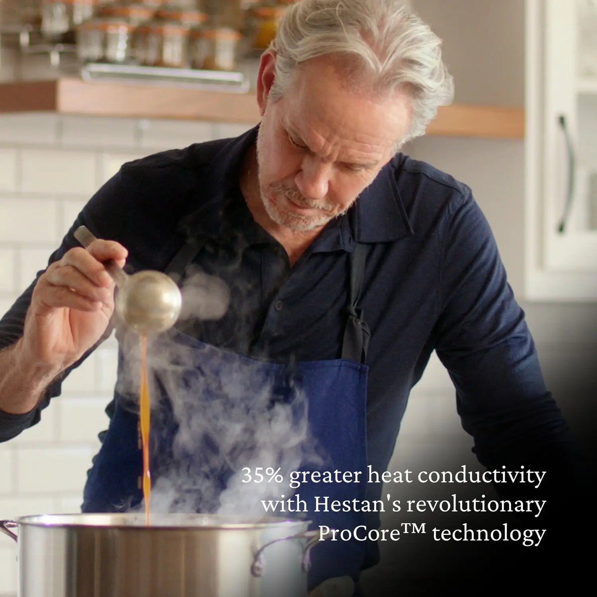 A man in a black shirt and blue apron ladles broth from a steaming pot in a modern kitchen. Text reads: 35% greater heat conductivity with the Thomas Keller Commercial Clad Stainless Steel 7-Piece Cookware Set.