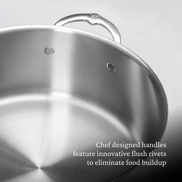 A close-up of the Thomas Keller Insignia Commercial Clad Stainless Steel Stock Pot features tri-ply construction and showcases flush rivets on the handle to prevent food buildup against a plain gray background. Text: Chef designed handles feature innovative flush rivets to eliminate food buildup.