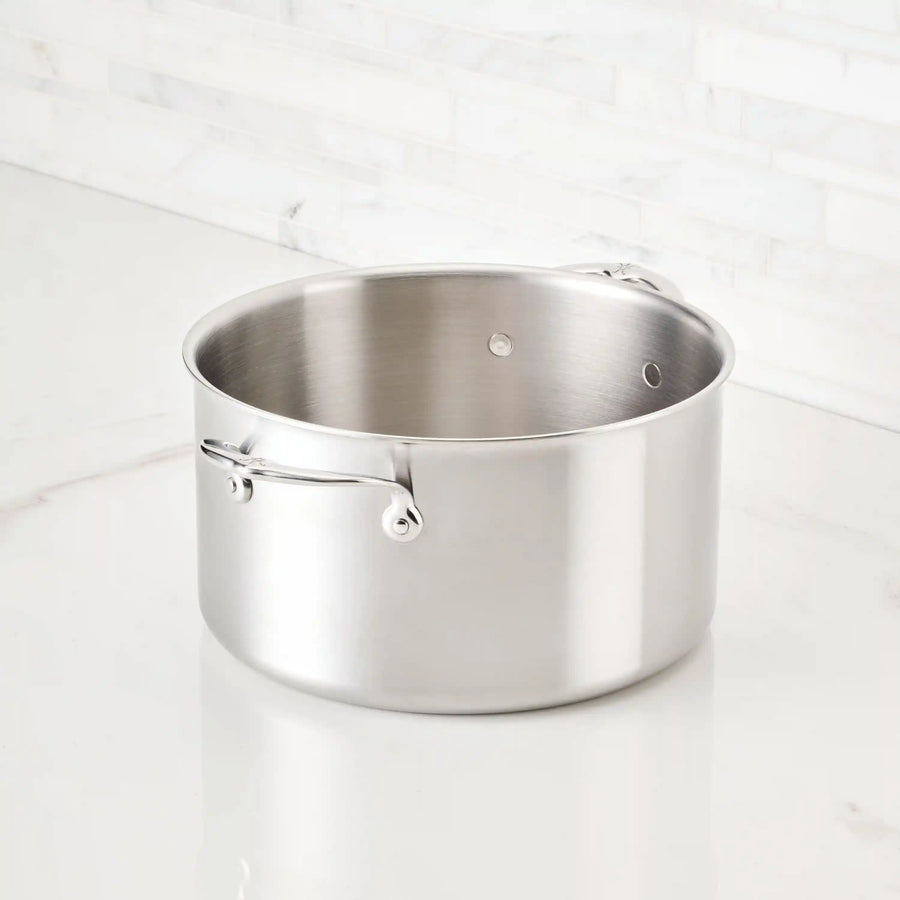 The Thomas Keller Insignia Commercial Clad Stainless Steel Stock Pot, featuring two side handles, sits on a white marble countertop against a tiled backsplash, ready to deliver professional culinary results.