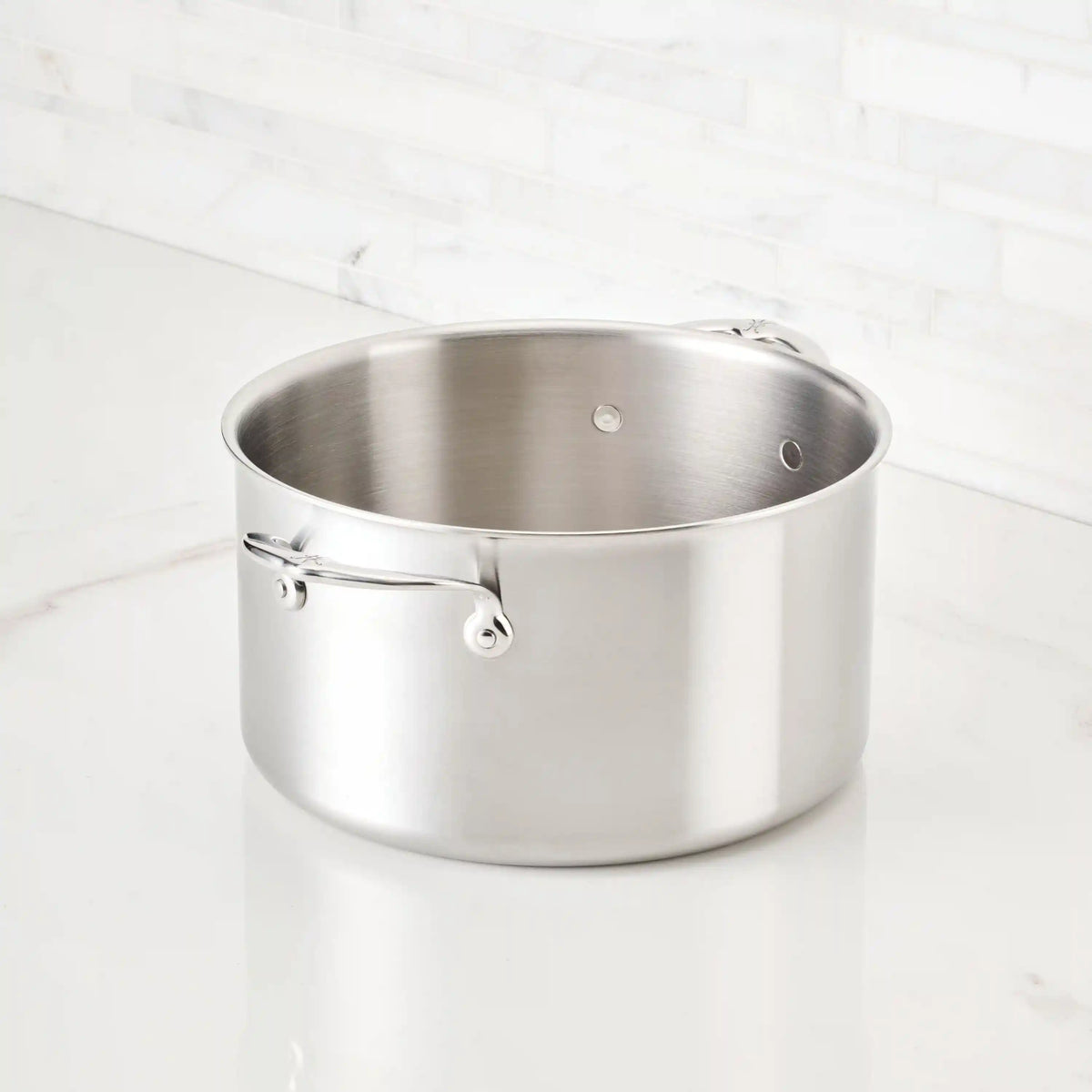 The Thomas Keller Commercial Clad Stainless Steel Stock Pot, with two handles, sits on a white marble countertop before a white tile backsplash, delivering professional-grade performance and sleek style.