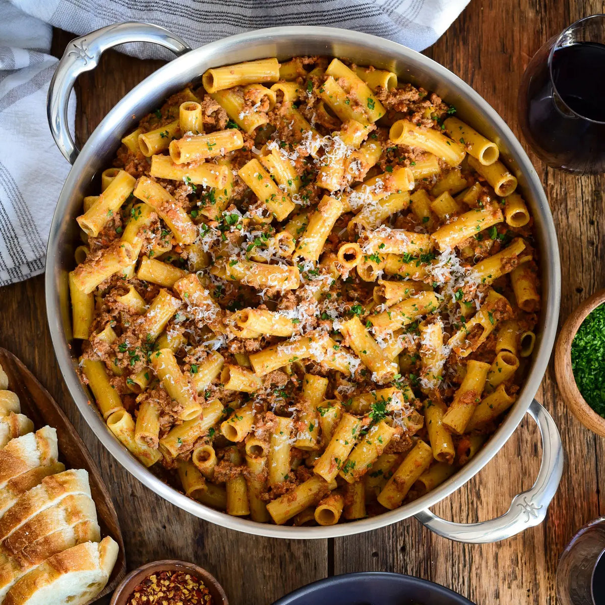 A large Thomas Keller Commercial Clad Stainless Steel Rondeau holds rigatoni pasta with meat sauce, grated cheese, and herbs on a wooden table next to red wine, sliced bread, parsley, and red pepper flakes.