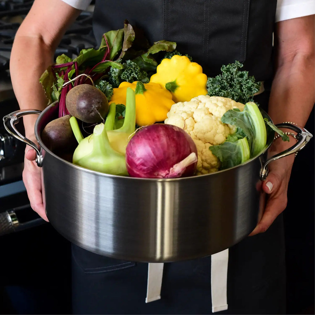 A person in a dark apron holds a large Thomas Keller Commercial Clad Stainless Steel Rondeau filled with fresh vegetables like beets, yellow squash, cauliflower, kohlrabi, red onion, and leafy greens.
