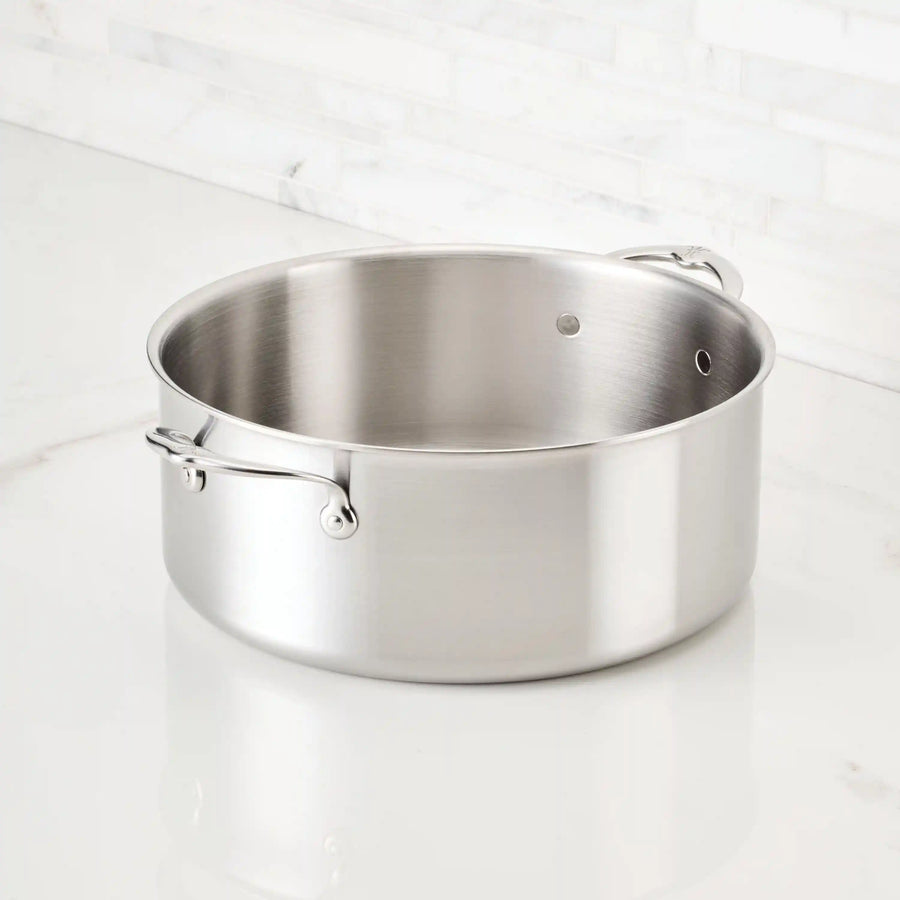 A Thomas Keller Insignia Commercial Clad Stainless Steel Rondeau with two handles and a brushed finish rests on a white marble countertop, lidless, against a backdrop of tiled white wall.
