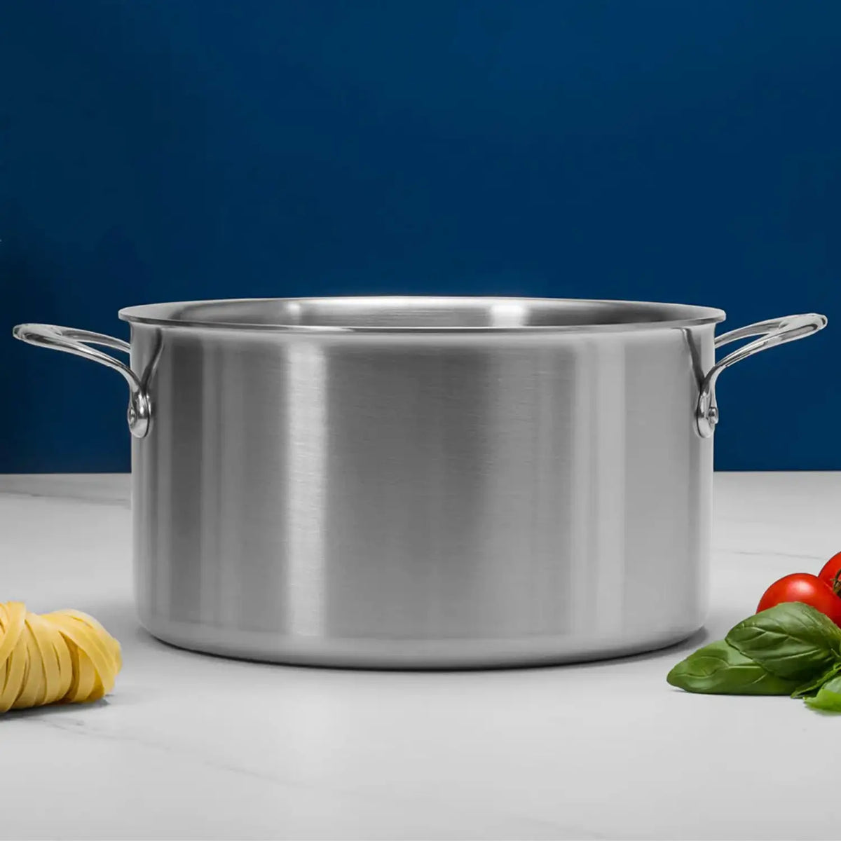The Thomas Keller Commercial Clad Stainless Steel Stock Pot, with dual handles, rests on a white surface. Uncooked pasta, basil, and tomatoes in the foreground evoke culinary expertise against a solid blue background.