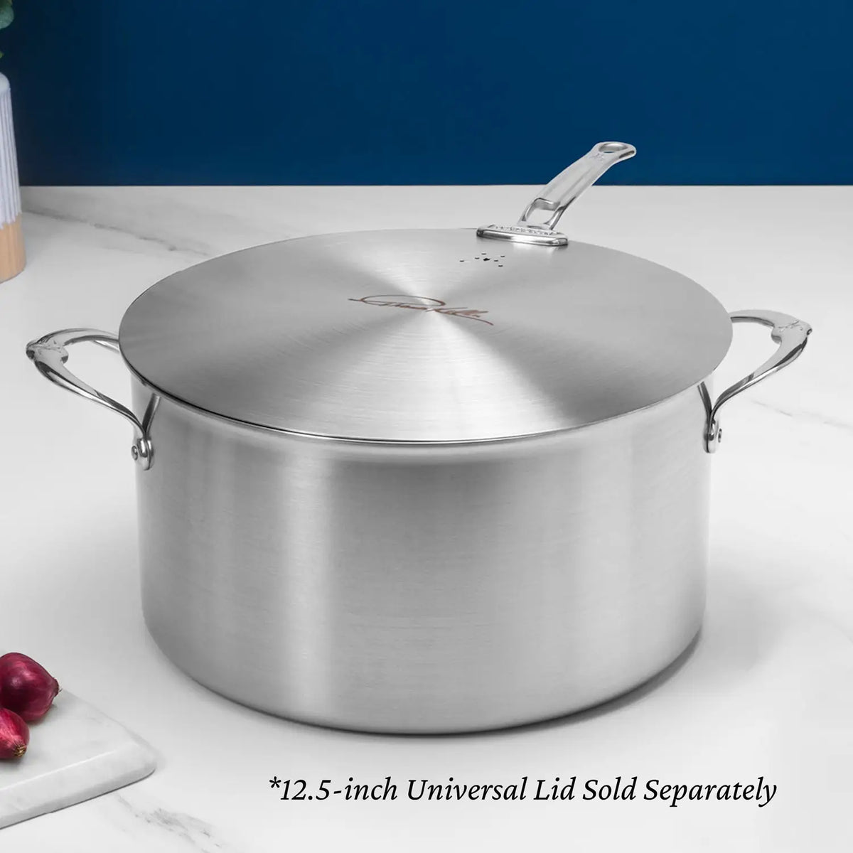 A Thomas Keller Commercial Clad Stainless Steel Stock Pot with two handles and a flat lid sits on a white countertop near three red onions. Text: 12.5-inch Universal Lid Sold Separately. Designed for professional culinary performance.