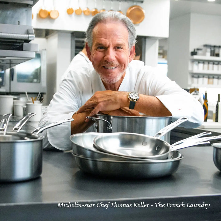 A chef in a white uniform leans on Thomas Keller Insignias Commercial Clad Stainless Steel Stock Pots in a commercial kitchen, with these tri-ply cookware pieces known for their excellent heat conductivity. The well-equipped kitchen showcases shelves and cooking utensils in the background.