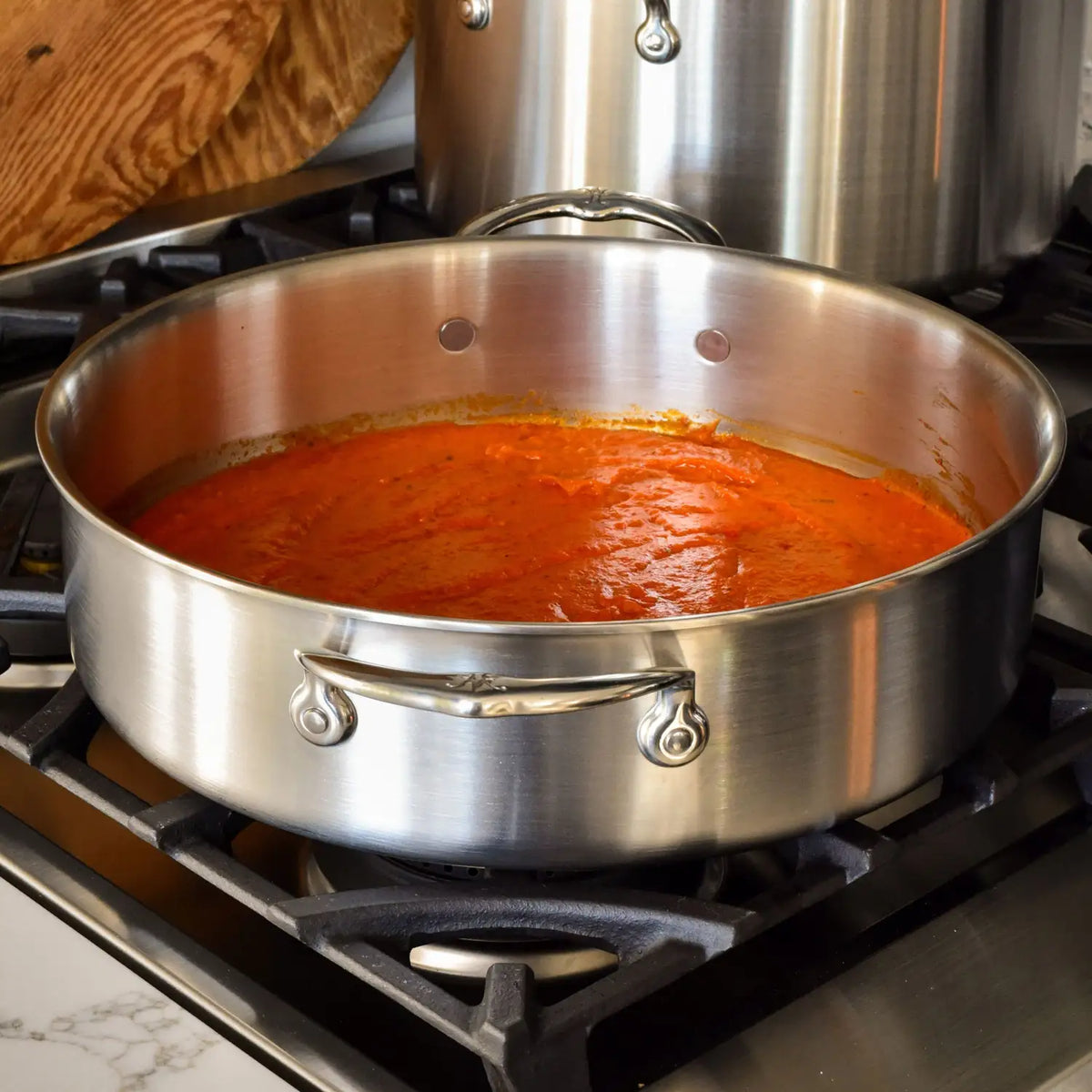 A Thomas Keller Commercial Clad Stainless Steel Rondeau filled with red tomato sauce sits on a stovetop burner, with a wooden cutting board and another pot visible in the background.