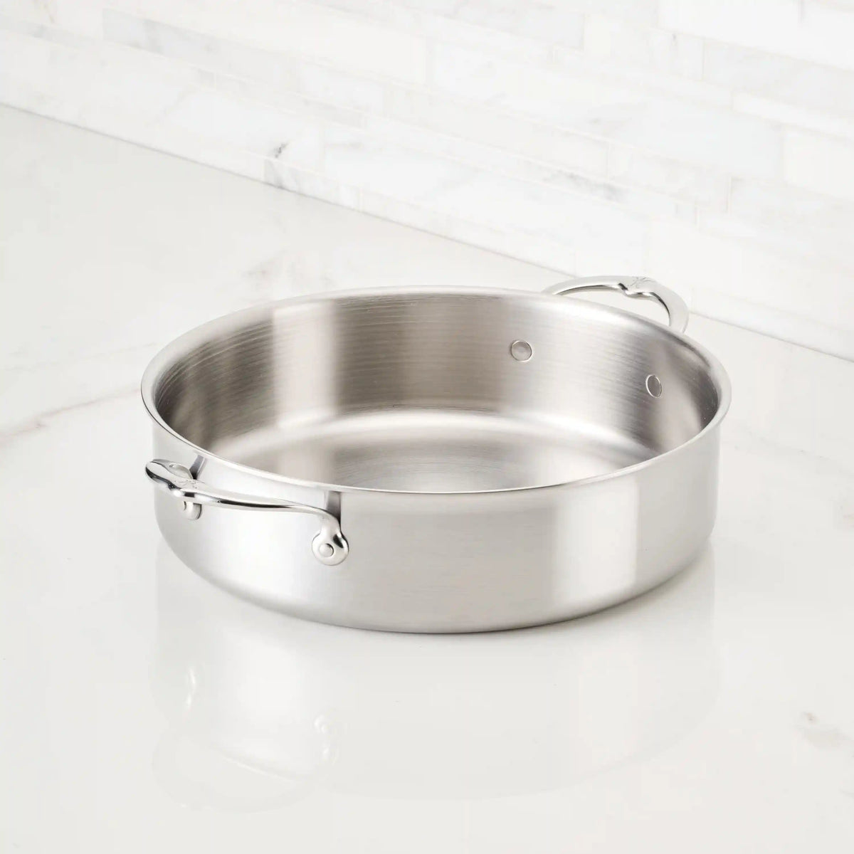 A Thomas Keller Commercial Clad Stainless Steel 11-Piece Cookware Set sauté pan with two handles and a brushed metal finish rests on a white marble countertop, set against a white tile backsplash.