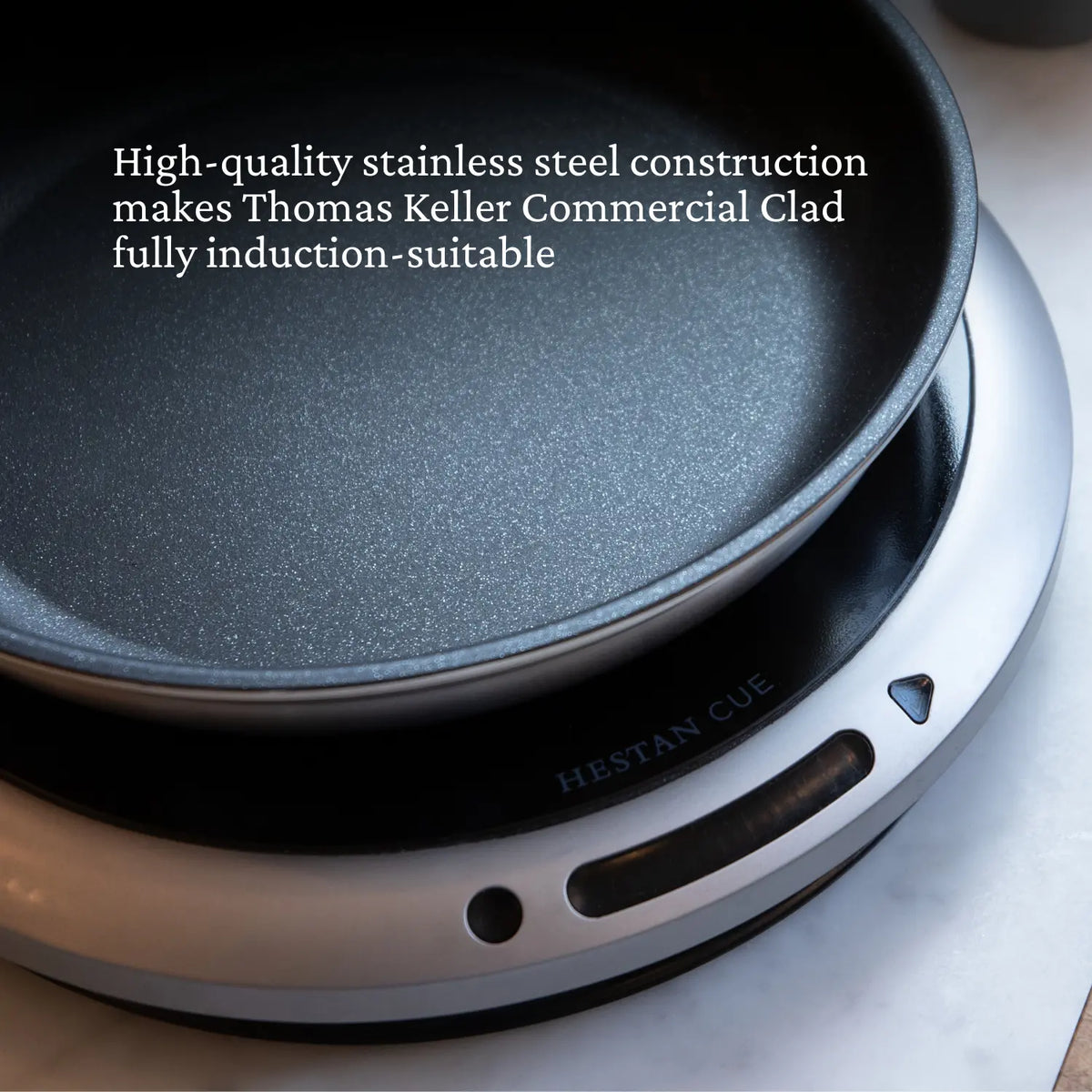 A close-up of a black Commercial Clad Stainless Steel TITUM® Nonstick Skillet by Thomas Keller Commercial Clad on a silver induction cooktop. Text emphasizes its professional performance and induction compatibility.