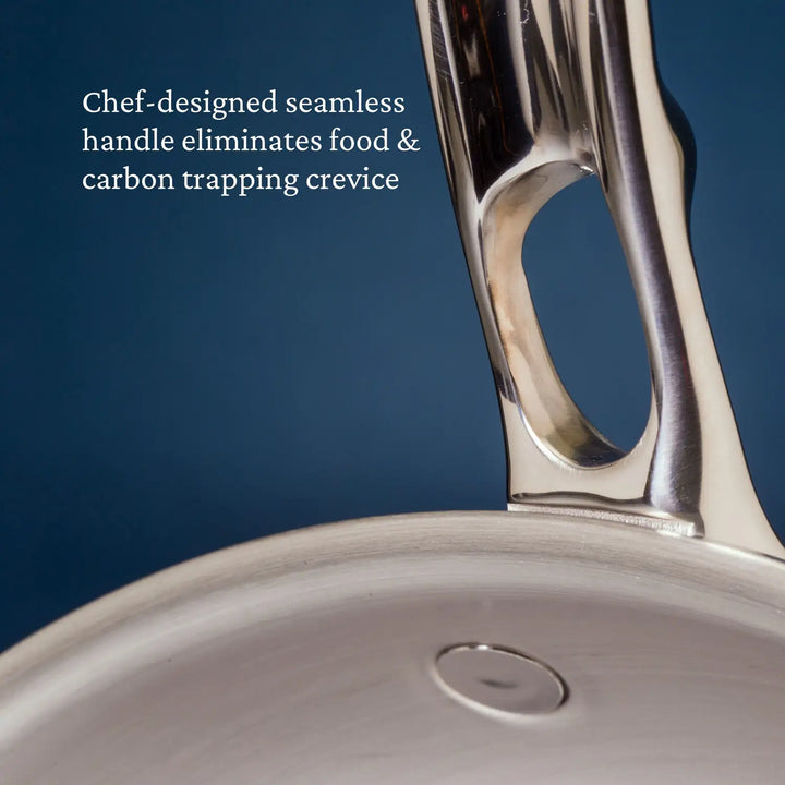 A close-up of the Thomas Keller Insignia Commercial Clad Stainless Steel Skillet features a seamless handle against a blue background. The text reads, Chef-designed tri-ply handle eliminates food & carbon trapping crevices and enhances heat conductivity.