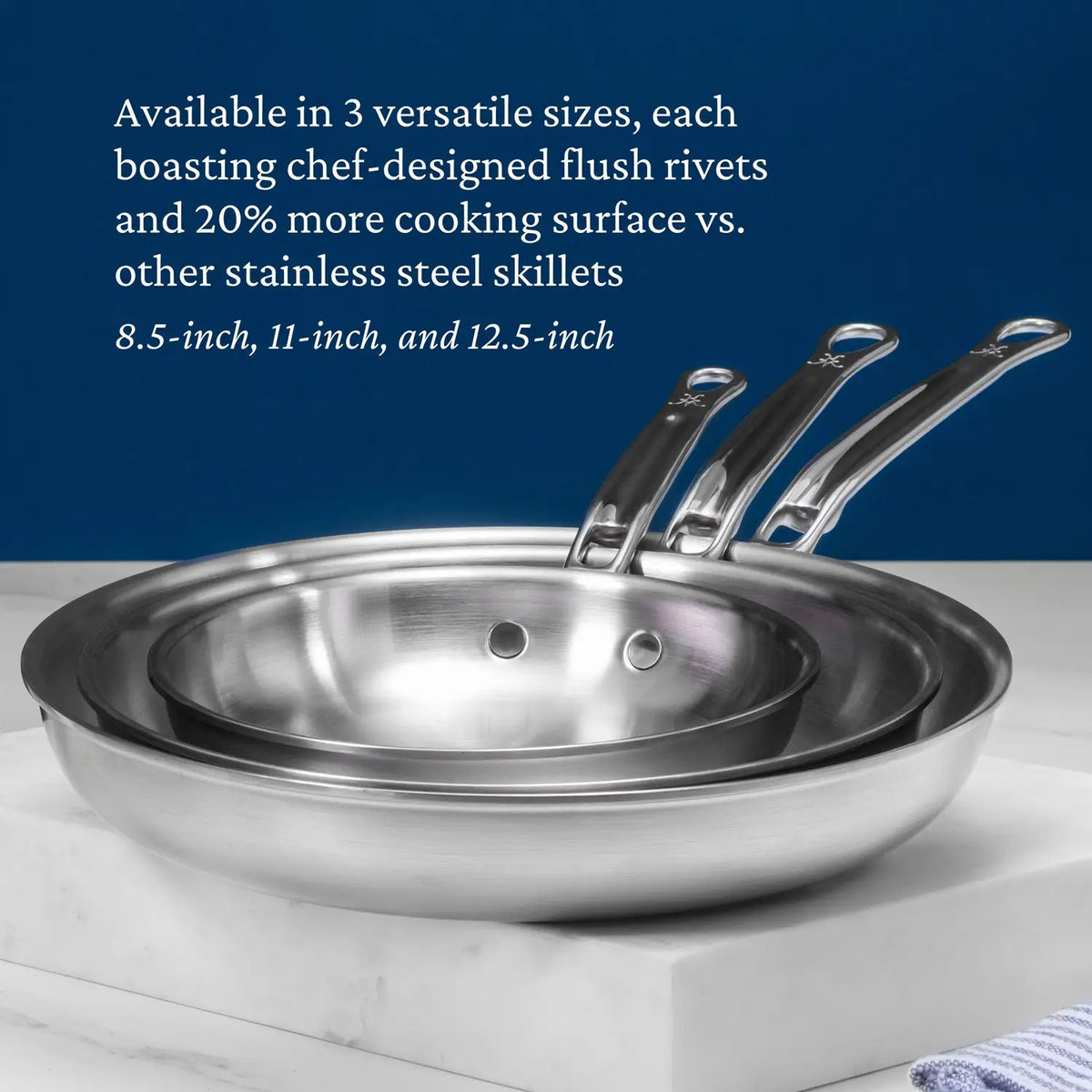 Three Thomas Keller Commercial Clad Stainless Steel Skillets (8.5”, 11”, 12.5”) are stacked on a gray surface, featuring flush rivets, increased cooking surface, and an eco-friendly design against a blue background.