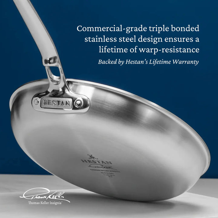 The Thomas Keller Commercial Clad Skillet is seen suspended off the kitchen counter, showing the side and underside of the skillet and its high quality stainless steel finish. Text on image reads: "Commercial-grade triple bonded stainless steel design ensures a lifetime of warp-resistance. Backed by Hestan’s Lifetime Warranty".