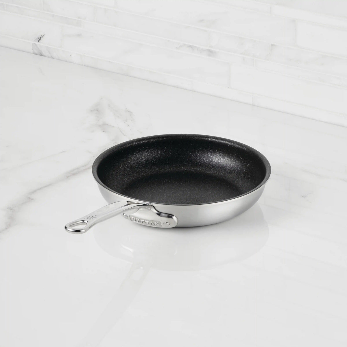 The Thomas Keller Commercial Clad Stainless Steel TITUM® Nonstick Skillet, with a sleek black interior, sits on a white marble countertop and backsplash, delivering professional-grade performance for any kitchen.