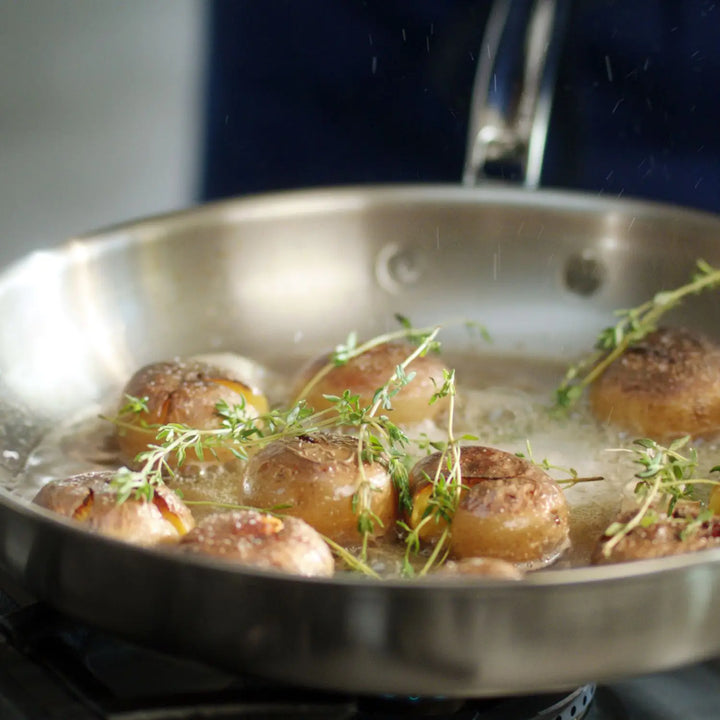 Mushrooms sizzle in a Thomas Keller Insignia Commercial Clad Stainless Steel Skillet with thyme, capturing perfect heat conductivity. Oil or butter melds the flavors seamlessly as the skillet rests on the stovetop.