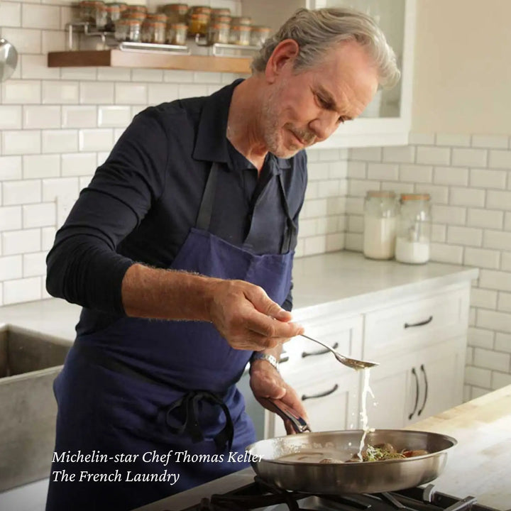 In a kitchen with white tiled walls and countertop jars, a man in a dark shirt and blue apron expertly cooks at the stove, demonstrating the heat conductivity of Thomas Keller Insignias Commercial Clad Stainless Steel Skillets as he adds liquid to the pan.