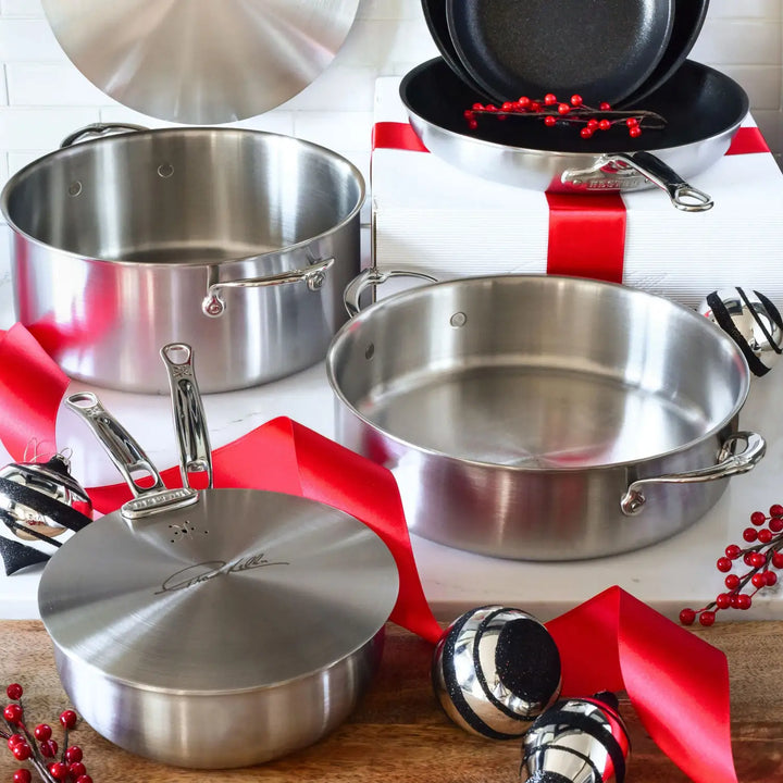 Thomas Keller Insignias Commercial Clad Stainless Steel Skillets, boasting exceptional heat conductivity, are elegantly displayed with red ribbons and ornaments. This cookware set rests on a wooden surface against a white background, featuring pots and pans with lids.