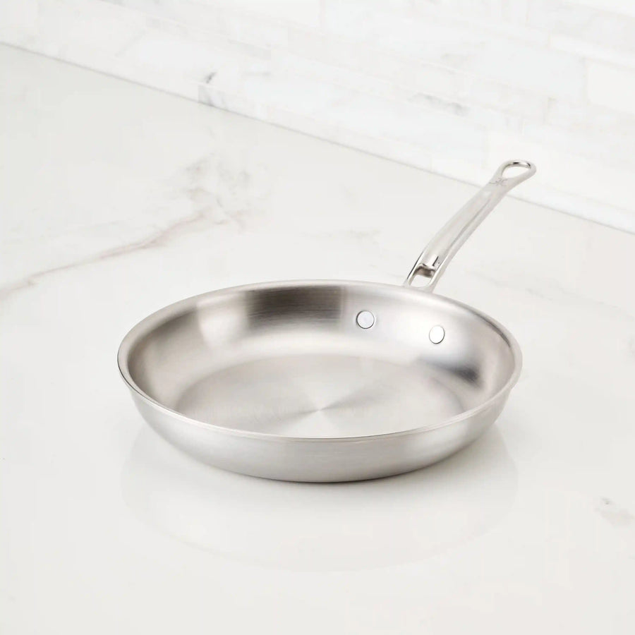 A Thomas Keller Insignia Commercial Clad Stainless Steel Skillet with a long handle rests on a white marble countertop. Featuring Hestan ProCore™ aluminum, it looks pristine and unused—perfect for professional culinary performance.