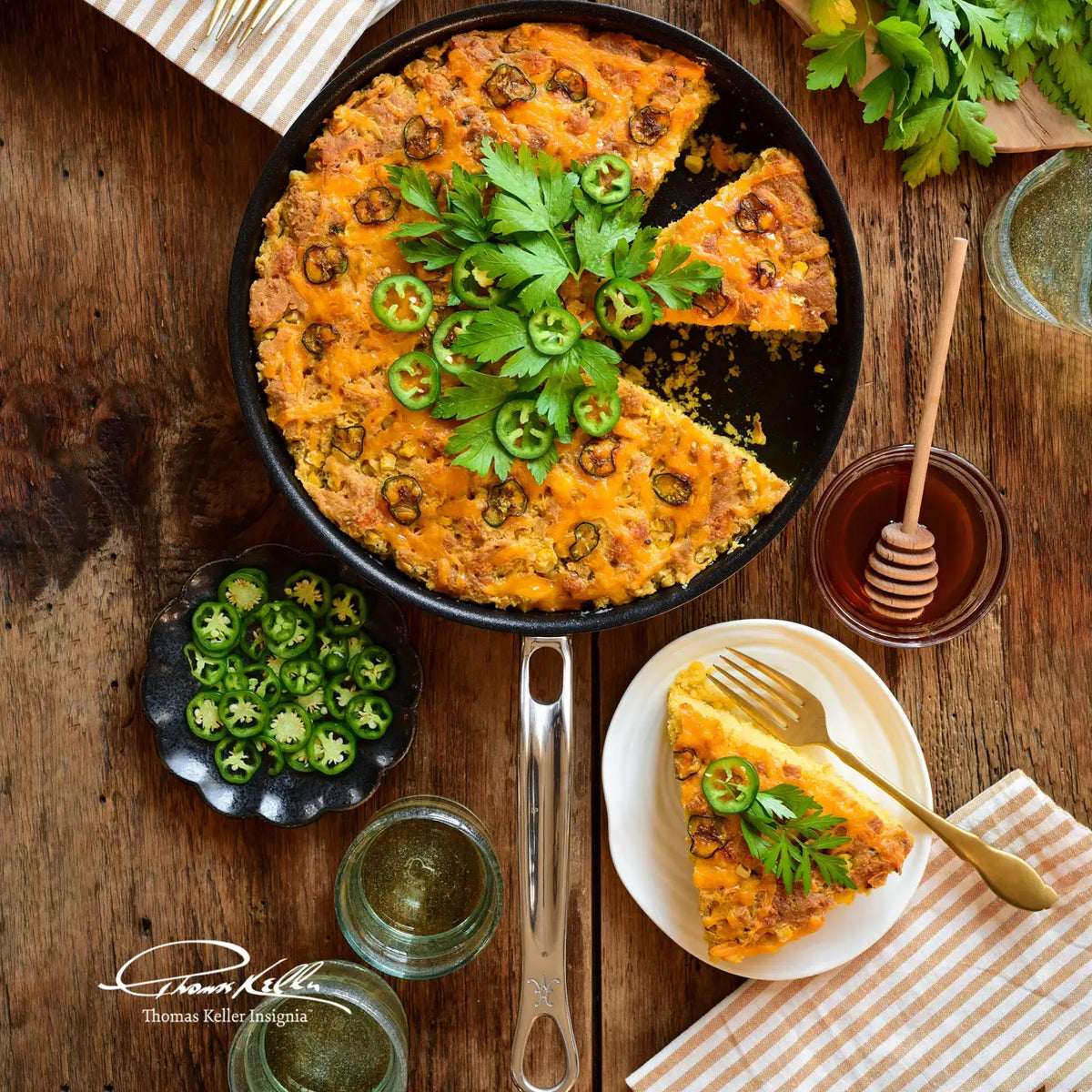 A skillet of cornbread topped with jalapenos and herbs sits on a table, baked in the Thomas Keller Commercial Clad Stainless Steel TITUM® Nonstick Skillet, with a slice plated beside glasses, honey, parsley, and sliced jalapenos.