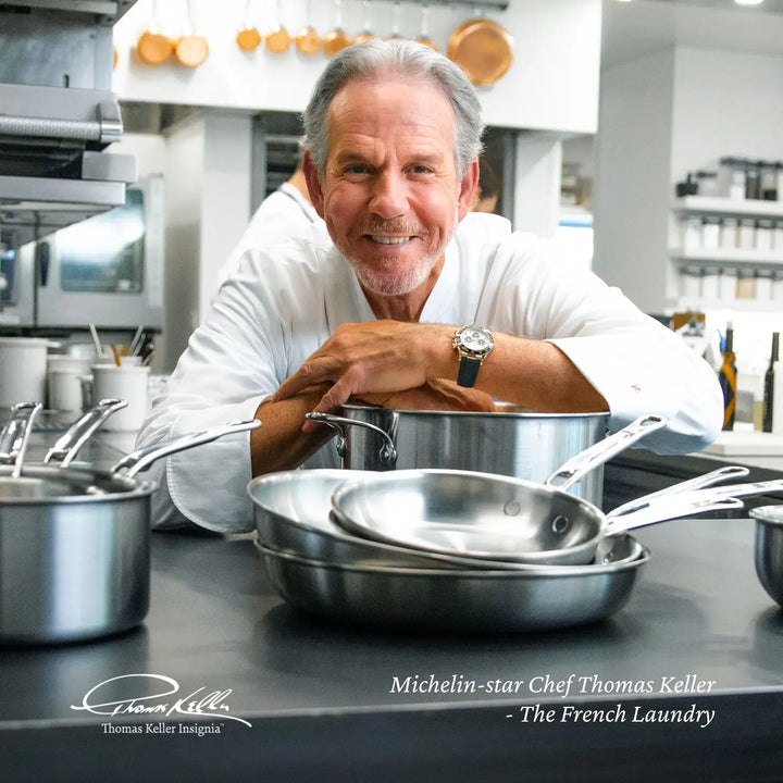 In a bustling kitchen, a chef in white is surrounded by glistening stainless steel cookware. Shelves full of essentials frame the scene, highlighting Michelin-star Chef Thomas Keller - The French Laundry, and showcasing the renowned Thomas Keller Insignia Commercial Clad Stainless Steel 7-Piece Cookware Set.